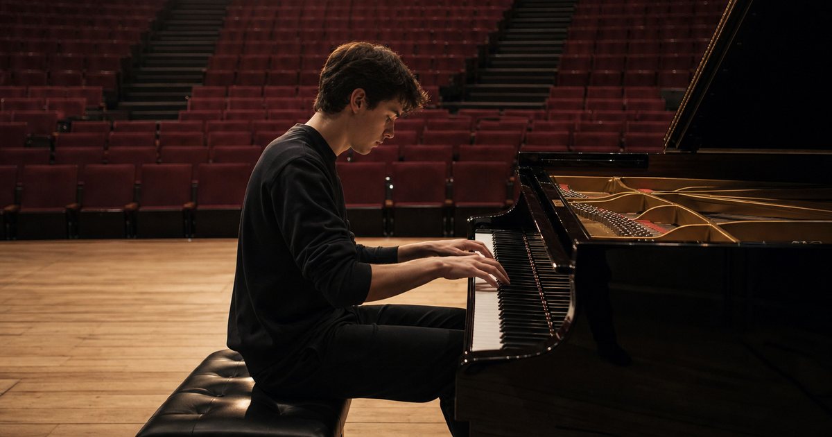 A young pianist in a concert hall during a rehearsal, alone on stage at a grand piano, empty red seats in the audience, a single stage light on