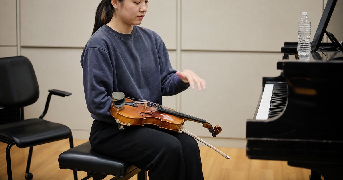 A young violinist shaking out her left hand during a break in a practice room, violin resting on her lap, a teacher's chair nearby and a water bottle on the piano